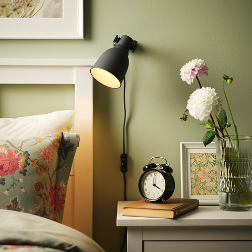 La lámpara de pared HEKTAR ilumina la mesita de noche con flores y reloj, junto a la cama con almohadas.