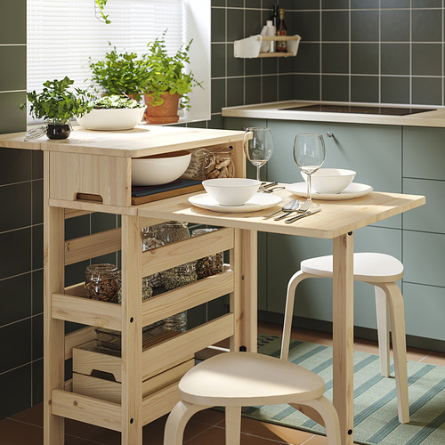 Cocina moderna con mesa de madera clara RESARÖ, dos taburetes, platos, vasos y macetas de hierbas sobre un fondo de pared con azulejos.