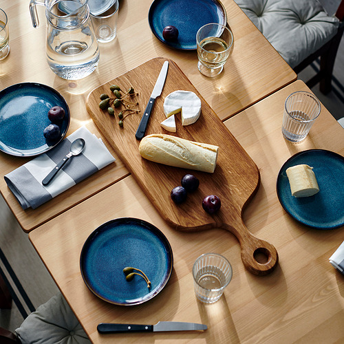 Tabla de embutidos de madera con quesos, pan, frutas y platos, vasos y servilletas azules.