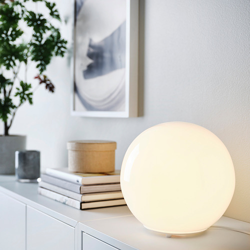 Lámpara de mesa FADO con pantalla grande y redonda de vidrio blanco en armario con plantas, libros, que emiten una luz suave.