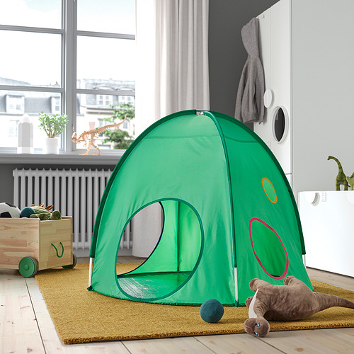 Los pequeños niños verdes juegan a la carpa, luminosa y aireada, colocada en el interior sobre una alfombra, con un juguete para perros cerca.