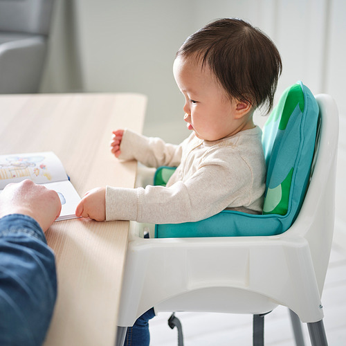 Un bebé sentado en una trona blanca con un cojín inflable azul verdoso como apoyo. El niño está tratando de alcanzar un libro sostenido por un adulto.