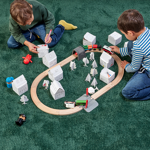Dos niños jugando con una maqueta de cartón MÅLA de una ciudad, una vía de tren y vehículos.