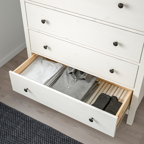 Cómoda de madera blanca con cuatro cajones, tres cerrados, uno abierto que deja ver ropa doblada y calcetines, anclado por seguridad.