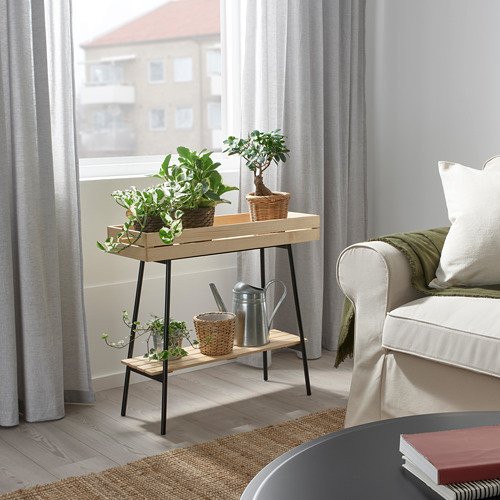 Soporte de plantas VANILJSTÅNG de tamaño real, de madera con patas negras, que sostiene varias plantas en macetas cerca de una gran ventana en un acogedor espacio habitable.