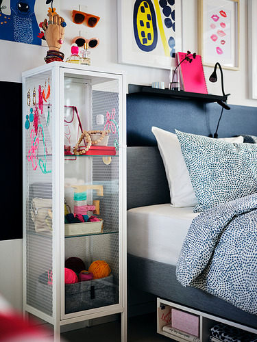 Rincón de dormitorio moderno con armario de almacenamiento BAGGEBO blanco que muestra accesorios coloridos. Cama gris con almohadas grandes, marcos de arte vibrantes en la pared.