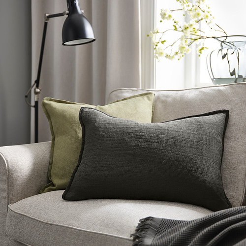 Acogedora sala de estar con almohada LAGERPOPPEL gris sobre sofá beige, lámpara negra y flores blancas.