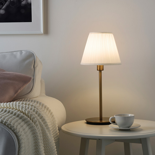 Acogedor rincón de salón con lámpara SKAFTET, sillón beige, taza de café y pared blanca. La lámpara es alta, delgada, de color latón, con una pantalla plisada blanca.