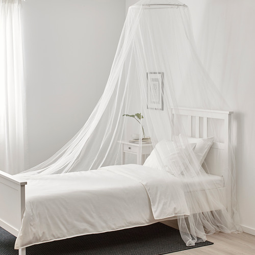 Cama con dosel blanco con red, estructura de plástico, funda de poliéster de fácil limpieza. Al lado de la mesita de noche con planta, cuadro, alfombra negra.
