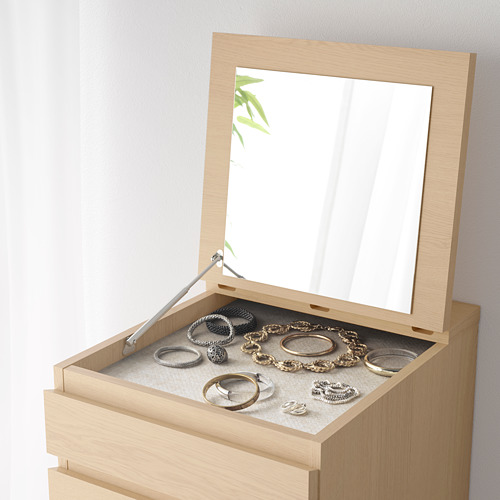 Una cómoda de madera clara con espejo y compartimento forrado de fieltro que contiene joyas y relojes.