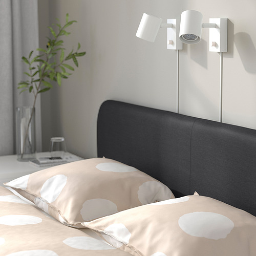 Escena de dormitorio con cama tapizada en negro contra pared blanca. La cama tiene dos luces montadas en la pared y una colcha beige estampada.