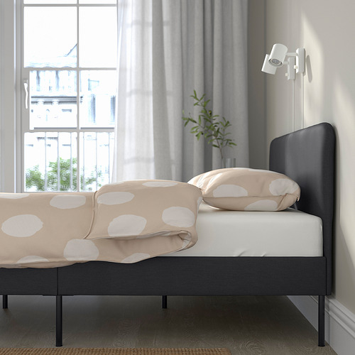 Dormitorio con estructura de cama SLATTUM, ropa de cama color canela, planta, ventana grande.