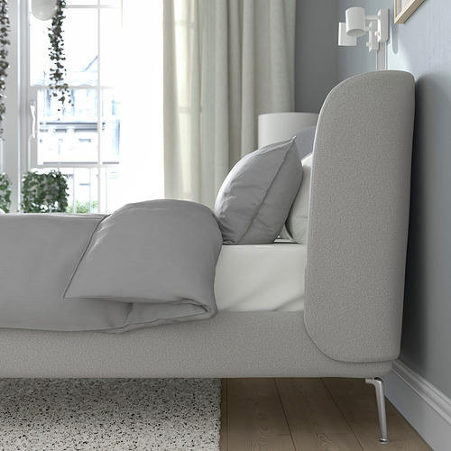 Cama moderna con cabecero curvo, tapizada en tela suave, ideal para relajarse. En la foto junto a un gran ventanal con luz natural.