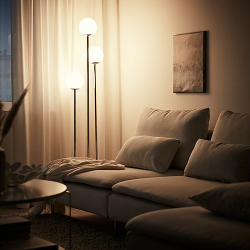 Acogedora sala de estar con sofá beige, lámpara de pie iluminada, cortinas y planta.