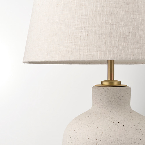 Lámpara de mesa moderna con una base crema texturizada, detalles de latón y una pantalla de tela blanca, que emana un brillo cálido y acogedor.