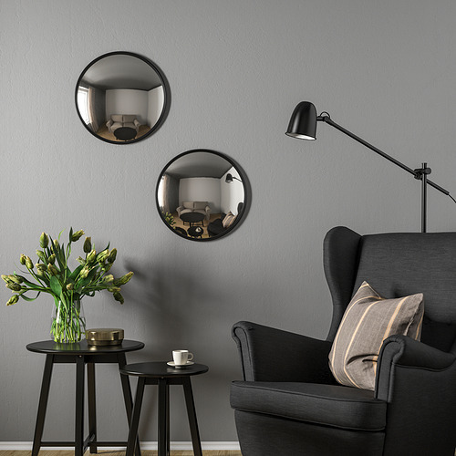 Dos espejos convexos en la pared gris reflejan el espacio, con sillón negro, lámpara y mesa floral debajo.