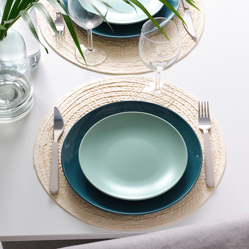 Mesa con platos verde azulado y manteles individuales tejidos. Se incluyen copas de vino, cubiertos y una planta.