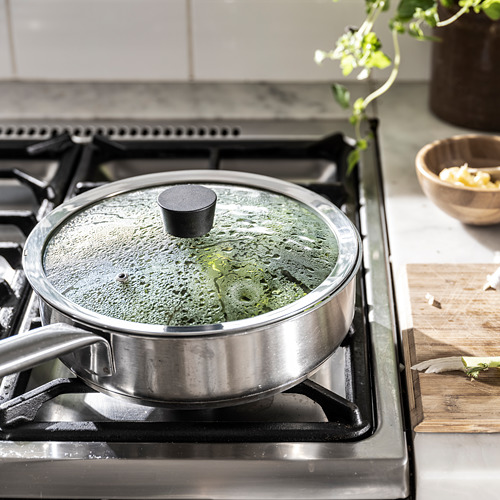 La sartén de acero inoxidable con tapa negra cocina las verduras, la tapa de vidrio muestra el progreso.