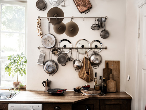 Pared de la cocina con ollas, sartenes y tapas para colgar organizadas en estantes cerca de una ventana. Mostrador de madera con tablas de cortar, plantas y utensilios debajo.