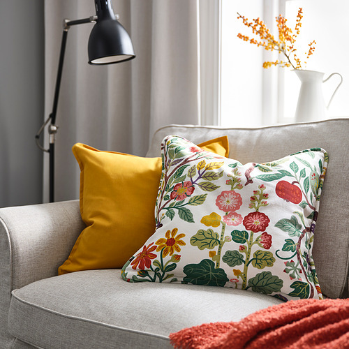 Acogedora silla con cojín floral VILDPERSILJA, almohada mostaza más grande, manta roja y flores naranjas en un jarrón blanco.