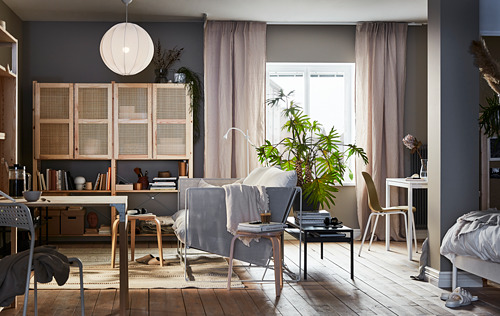 Salón moderno con estanterías de madera IVAR, paredes grises, luz colgante, cortinas beige, plantas y muebles minimalistas.