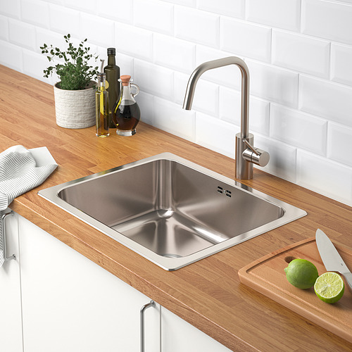 Fregadero de cocina de acero inoxidable con grifo sobre encimera de madera. Lime y cuchillo en la tabla de cortar, botellas y planta cercana.