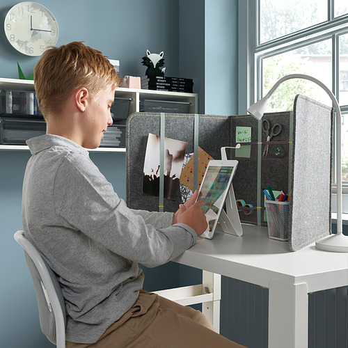 Niño sentado en un escritorio blanco usando una tableta. El escritorio tiene un divisor gris para privacidad y amortiguación del sonido; Contiene notas y suministros.