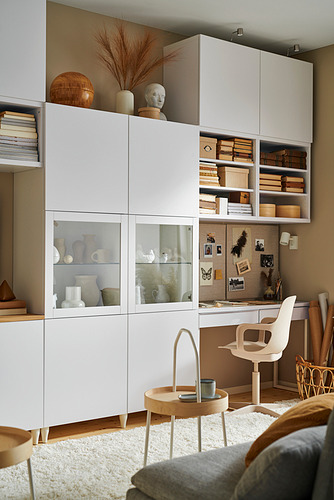 Moderna oficina en casa con armarios blancos BESTÅ, puertas de cristal, libros, artículos de decoración, silla y mesita con taza.