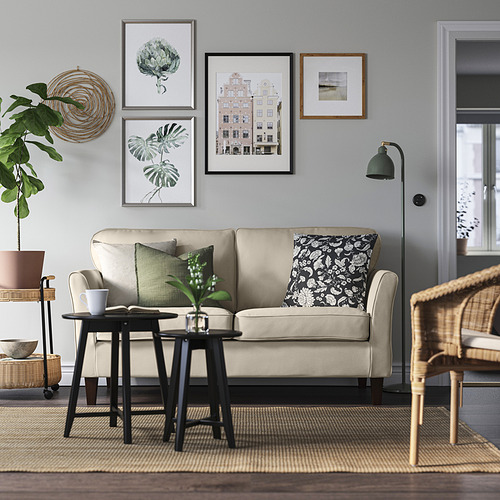 Salón minimalista moderno: sofá EKHOLMA beige, mesa auxiliar negra, silla de mimbre, alfombra gris, plantas verdes, arte mural.