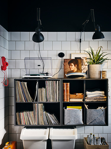 Mueble de almacenamiento de pared EKET negro con estantes y contenedores, con capacidad para discos, libros y decoración, con luces industriales en la parte superior.