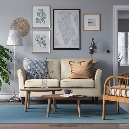 Sofá EKHOLMA beige, patas de madera, pared de galería, alfombra azul, silla de mimbre, mesa de centro, lámpara: elegante sala de estar.