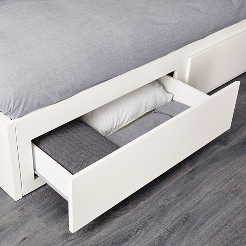 La imagen muestra una cama de almacenamiento blanca con un cajón abierto, revelando almohadas y ropa de cama.