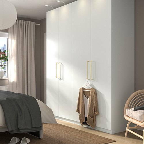 Dormitorio moderno con armario PAX blanco, manijas doradas, suéter, cama de lino oscuro / claro, silla de mimbre cerca de cortinas transparentes.