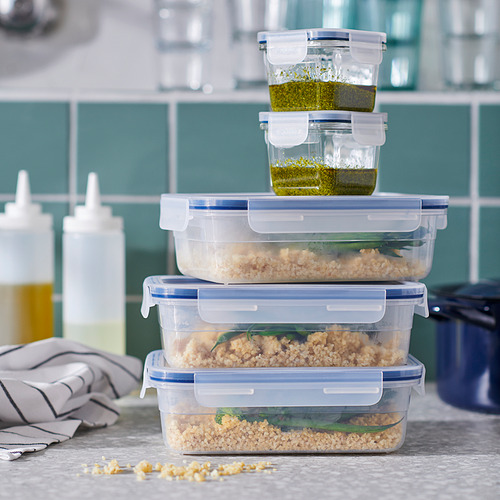 Recipientes de plástico transparente para alimentos apilados con tapas azules en una encimera. Diferentes tamaños, sobras almacenadas, salsa verde encima.