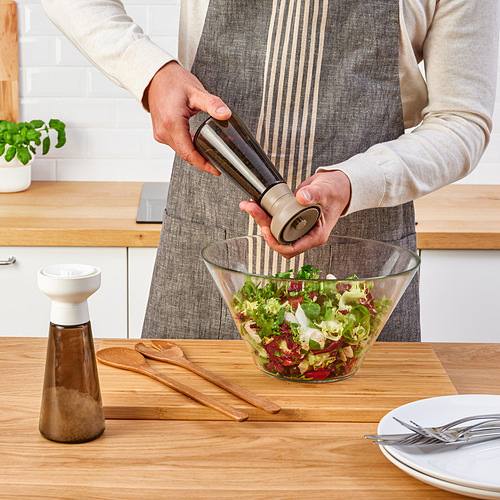 Una persona muele pimienta en una ensaladera con un molinillo de especias HALVTOM sobre una mesa de madera.