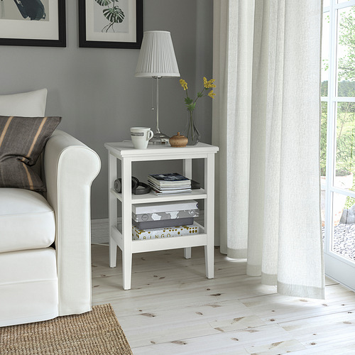 Pequeña mesa auxiliar blanca de IDANÄS junto al sofá, con lámpara, tazas y libros. Suelo de madera clara, pared gris, cortinas transparentes.