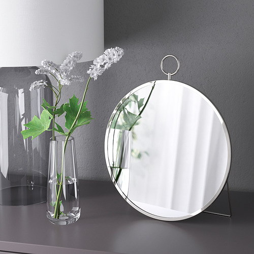 Un espejo redondo en un soporte junto a un jarrón transparente con flores blancas. El espejo puede colgar o estar de pie.