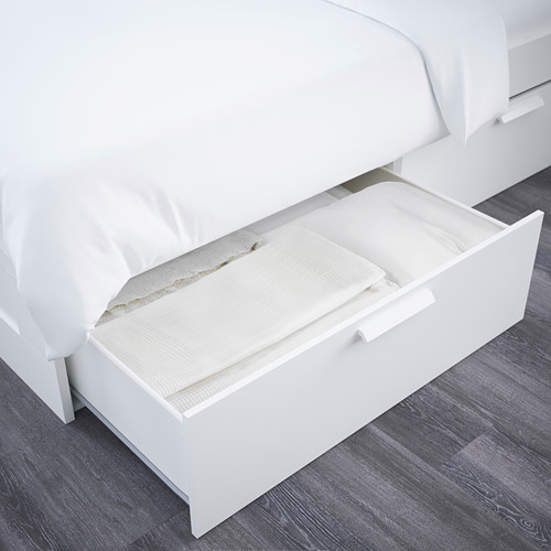 Esta imagen muestra un cajón de almacenamiento blanco abierto debajo de la cama, lleno de ropa de cama blanca cuidadosamente dispuesta.