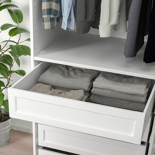 Cajón blanco de pax abierto, ropa gris cuidadosamente doblada en el interior. Planta en suelo de madera al lado.