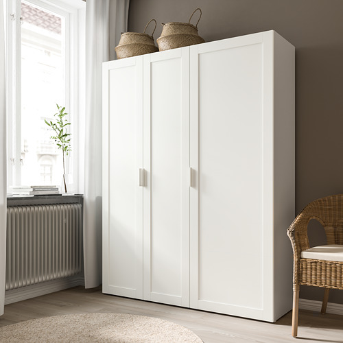 Moderno armario blanco con dos puertas y cestas tejidas en la parte superior, junto a una ventana con radiador y planta.