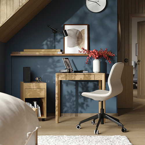 Moderna oficina en casa con escritorio de madera clara, silla blanca, computadora portátil, lámpara, libros y flores rojas.