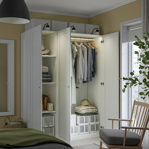 Armario blanco abierto que muestra ropa, toallas dobladas y cajas de almacenamiento en una habitación con una silla verde y una ventana grande.