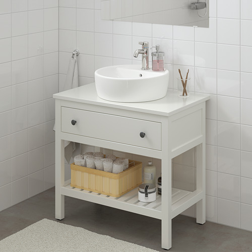 Tocador HEMNES blanco con lavabo redondo, cajón y estante. La cesta contiene papel higiénico y champú, toalla gris cerca.