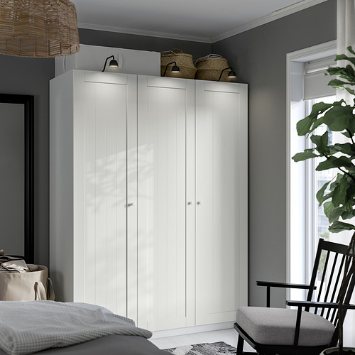 Un armario blanco PAX con armarios elegantes y modernos y un diseño minimalista. Se mantiene erguido, ocupando gran parte del espacio, con cestas tejidas en la parte superior.