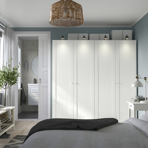 Un dormitorio moderno, un gran armario blanco PAX contra una pared verde azulado, resaltado por una iluminación cálida y detalles en negro.