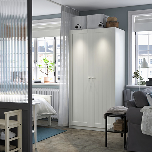 Habitación moderna con armario PAX blanco, alto y estrecho, encaje entre ventanas. Sofá gris y alfombra azul, paredes grises.