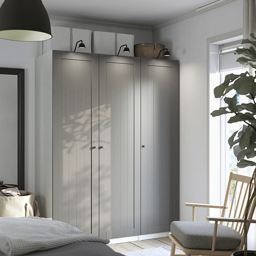 Un dormitorio moderno con un gran armario PAX gris, alto y elegante, apoyado en la pared, flanqueado por una lámpara colgante y una pequeña silla de madera.