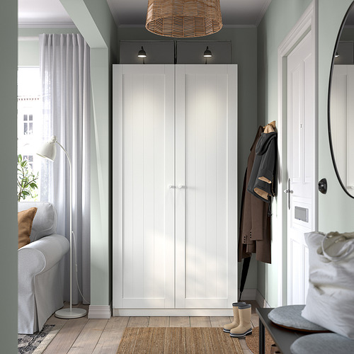 Pasillo moderno con armario alto blanco PAX, abrigos, botas, sofá gris claro y gran ventana con cortinas transparentes.