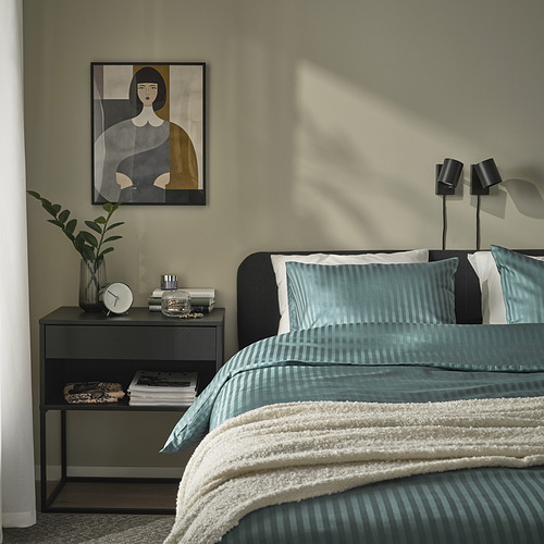 Dormitorio minimalista con edredón NATTJASMIN azul verdoso, rayas sutiles, mesita de noche con reloj y libros.