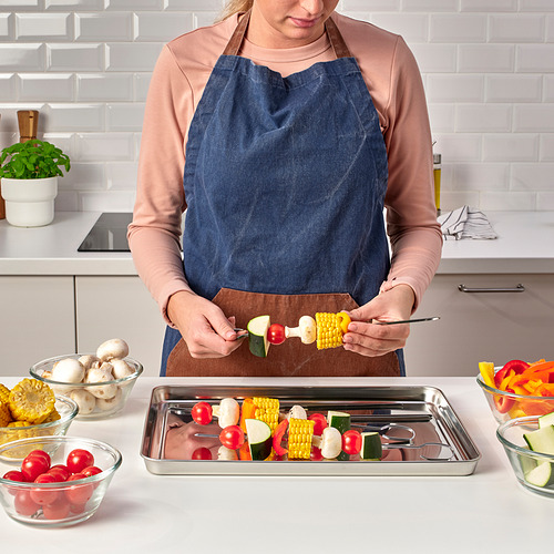 Una persona prepara brochetas, con verduras de colores y maíz en una bandeja metálica de la serie GRILLTIDER, ideal para barbacoas.